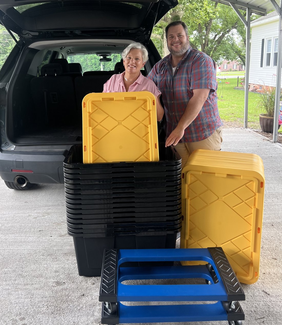 Drew and Crystal, owners of Hometown Tote Rental, standing behind their SUV with a stack of totes, a dolly, and a lid ready to load